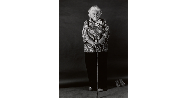 A black-and-white portrait photograph, in full length, of a white-haired, elderly White woman wearing a plaid shirt, long dark pants, and resting her hands on a cane.