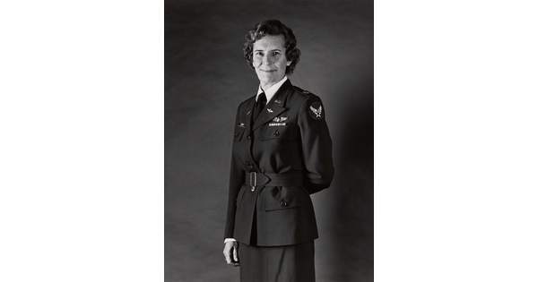 A black-and-white portrait photograph, three-quarter length, of a middle-aged White woman wearing a belted military jacket, one arm resting behind her back.