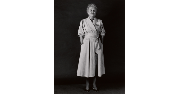 A black-and-white portrait photograph of an older White woman with short, light hair wearing a wrap dress with a name badge above her breast and her hands in her pockets.