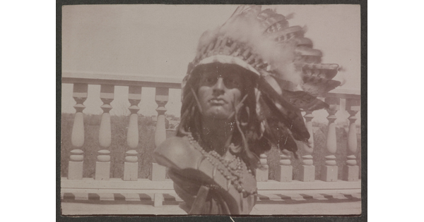 A sepia-toned photograph of a male Native American sculpture bust wearing a real feathered headdress and beads around the neck.