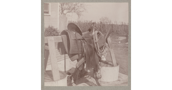 A sepia-toned photograph of the side view of a saddle and rope resting on a wooden sawhorse in a yard.