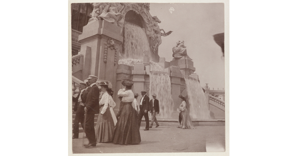 A sepia-toned photograph of women in long skirts and men in hats and jackets strolling by an elaborately sculpted facade with waterfalls.