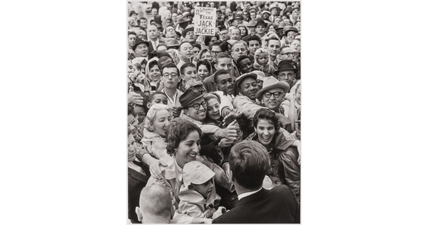 A black-and-white photograph of President Kennedy from the back facing a large crowd that is reaching up to touch his hand.