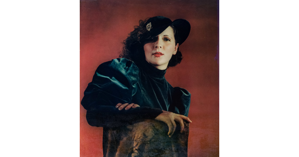 A color portrait photograph of a White woman with curly hair topped with a hat that covers one eye, wearing a green velvet dress with puffy sleeves, one arm resting on the back of a chair.