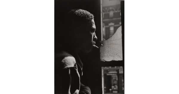 A black-and-white photograph of a Black man in profile with a cigarette in his mouth looking out of a shattered window.
