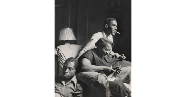 A black-and-white photograph of a Black woman knitting in an armchair, a young Black man smoking on the arm of her chair, and a young Black man seated on the floor reading.
