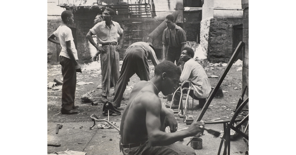 A black-and-white photograph of group of Black men talking and working in a rundown space littered with debris.