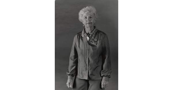 A black-and-white portrait photograph of an older White woman with short, curly gray hair wearing a long-sleeved button-down blouse.
