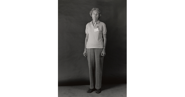 A black-and-white portrait photograph of an older White woman wearing a short sleeved polo shirt with a nametag pinned to her breast, and plaid trousers.