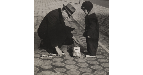 A black-and-white photograph of a small boy bundled in winter clothing watching as a man in a fedora crouches down to reach toward a toy fire truck at the boy&#039;s feet.