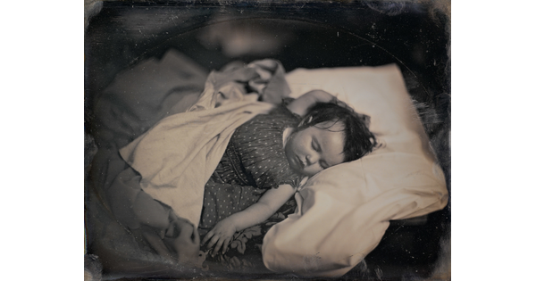A black-and-white photograph of a White baby sleeping on their back, dark hair sticking to their head, in a bed with one hand above their head and the other at their side.