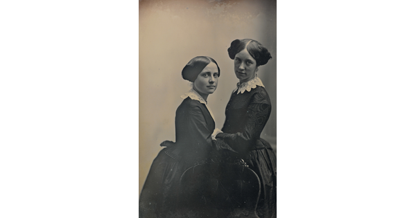 A black-and-white portrait photograph of two young White girls facing each another in period clothing and hairstyles.