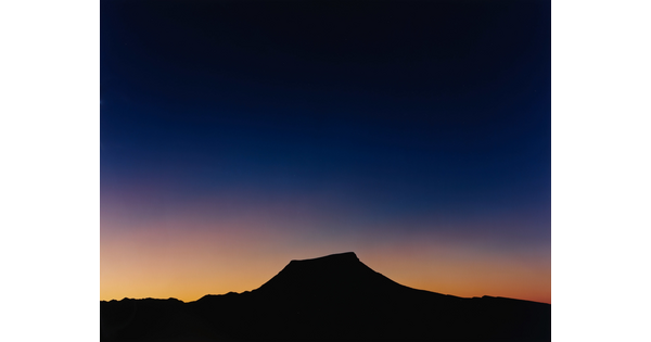 A color photograph of a sky that goes from almost black to blue to gold where it meets the silhouette of a flat topped mountain.