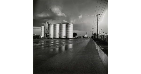 A black-and-white photograph of four grain elevators to the left of a wet road, a water tower in the far distance, and a flash of lighting behind power lines on the right.