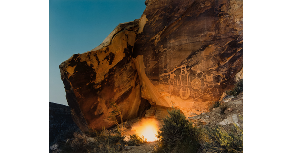 A color photograph of small campfire illuminating a rock wall with several human-like petroglyphs on it.