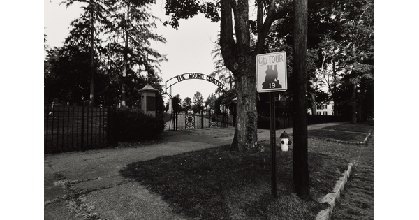 A black-and-white photograph taken from the street of a wrought iron gate at the entrance to a cemetery.