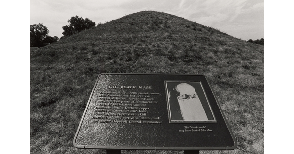 A black-and-white photograph of an outdoor informational plaque titled "The Death Mask" in front of a large grassy mound.