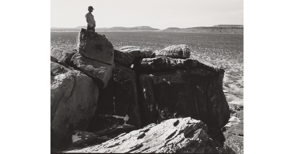 A black-and-white photograph of a man standing on a rocky cliff overlooking a wide canyon.
