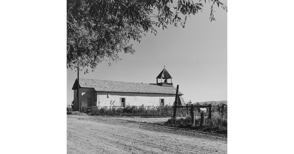 A black-and-white photograph of a long, narrow white building with a small steeple on a dusty road behind a fence.