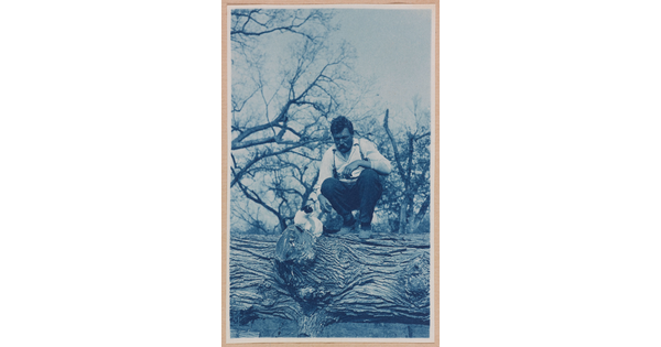 A black-and-white photograph of a White man squatting on a large log next to a white-and-black cat.