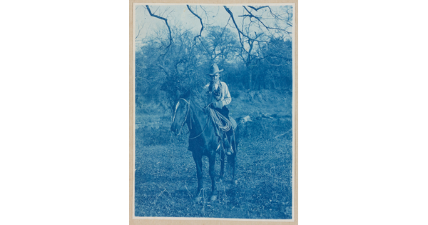 A black-and-white photograph of a man wearing a cowboy hat on horseback, pointing his revolver toward the viewer.