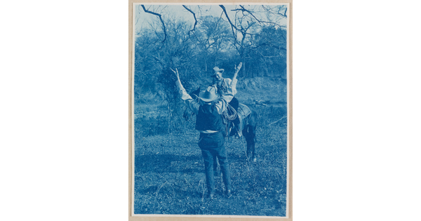 A black-and-white photograph of a man in a cowboy hat standing with both hands raised up in front of a smiling man wearing a cowboy hat on horseback.