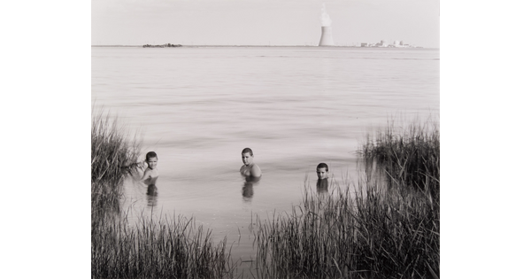 A black-and-white photograph of three White boys submerged to their shoulders in a lake; on the opposite shore in the background, a nuclear reactor puffs steam into the sky.