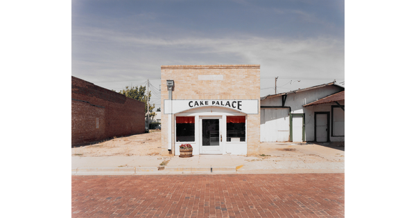 A color photograph of a tan brick building with a sign that reads "Cake Palace" above the door on a red brick road.