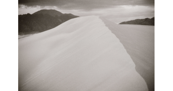 A black-and-white landscape photograph of the ridge of a sand dune against dark mountains and cloudy sky.