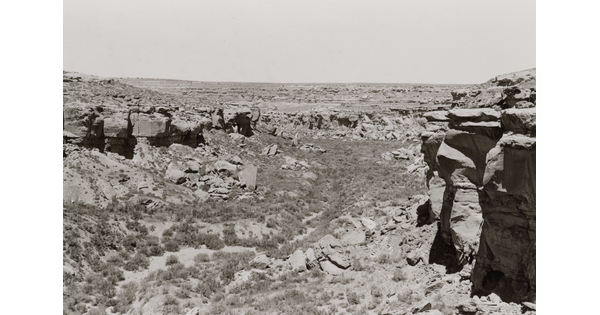 A black-and-white photograph of a scrubby desert landscape framed by high rocks and cliffs.
