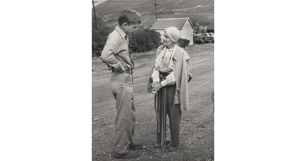 A black-and-white photograph of a White man and a White woman talking to each other on a gravel road.