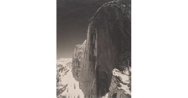 A black-and-white photograph of a steep cliff face and the snow-covered mountains around it.