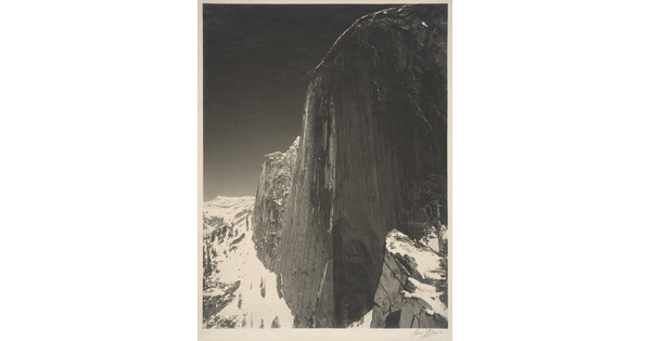 A black-and-white photograph of a steep cliff face and the snow-covered mountains around it.