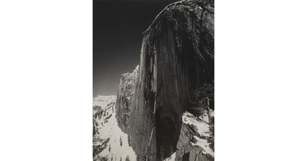 A black-and-white photograph of a steep cliff face and the snow-covered mountains around it.