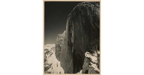 A black-and-white photograph of a steep cliff face and the snow-covered mountains around it.