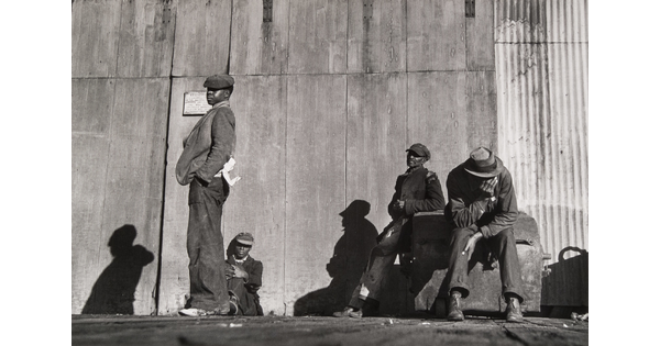 A black-and-white photograph of four Black men, three sitting and one standing, all wearing hats in front of a corrugated metal wall.