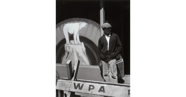 A black-and-white photograph of a Black man in a flat cap and dark cardigan behind a wooden barricade that says "WPA" and in front of a sign with a painting of a polar bear.