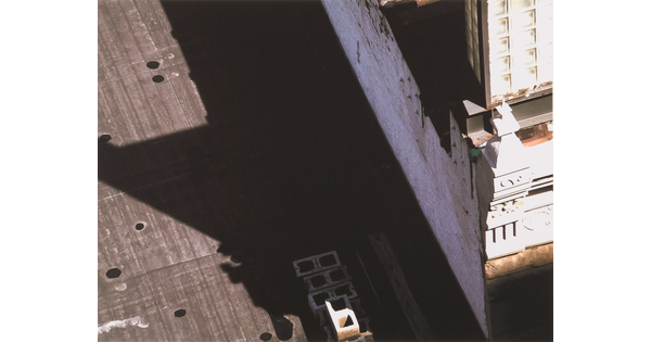 A black-and-white photograph of a building and the shadow it casts across the top of a neighboring building as viewed from the above.