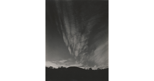 A black-and-white photograph of hills and trees silhouetted against a sky with thin, vertical clouds.