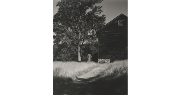 A black-and-white photograph of a person walking on a grassy path that leads to large barn next to a tree.