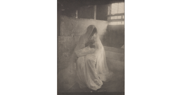 A soft-focus, black-and-white photograph of a seated woman wearing a white dress and veil, holding a baby wrapped in a white blanket.
