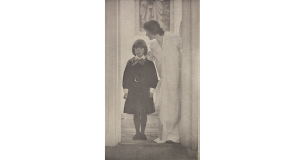 A soft-focus, black-and-white photograph of a White woman in a long, white dress with her arm around the shoulders of a White girl wearing a dark dress standing in a door frame.