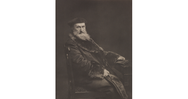 A black-and-white portrait photograph of a seated older White man with a long beard, wearing a beret and a long, fur-lined coat, holding a book in one hand.