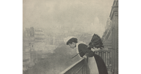 A black-and-white photograph of a White woman dressed as a maid in a white cap, black dress, and white pinafore holding a feather duster under her arm as she peers over a balcony at a smoggy cityscape.