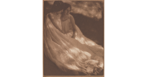 A sepia-toned photograph of two White women, both wearing long flowy dresses, sitting close together on the ground.