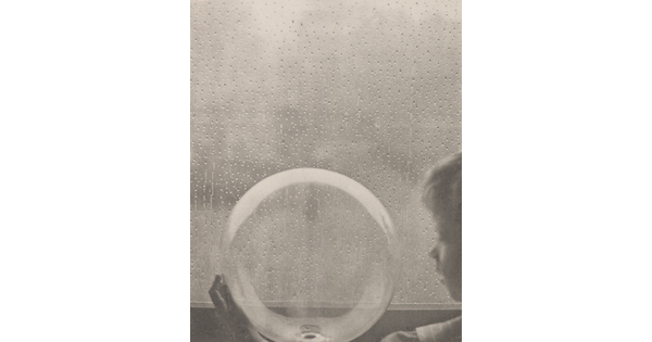 A black-and-white photograph of a White child looking at a clear glass globe in front of a rain-spattered window.