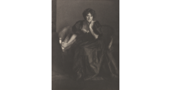 A black-and-white portrait photograph of a White woman, hair in an updo, wearing a long formal dress with puffy sleeves seated on a small sofa.