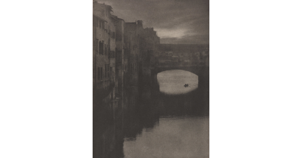 A sepia-toned photograph of a wall of tall buildings lining a river and an enclosed arched bridge stretching over the water.