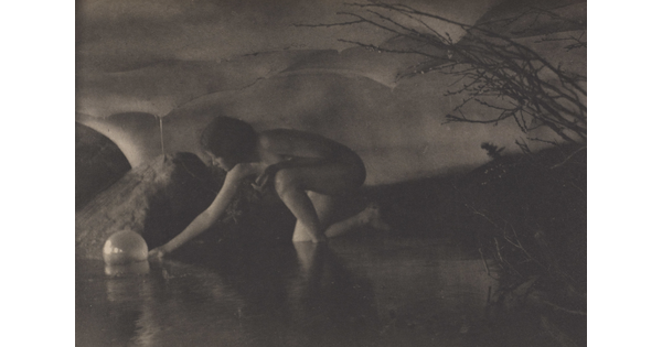 A black-and-white photograph of a nude White women reaching down to pick up a sphere in a shallow body of water.