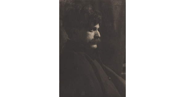 A black-and-white photograph of a White man with lots of dark, curly hair and a mustache sitting in profile.
